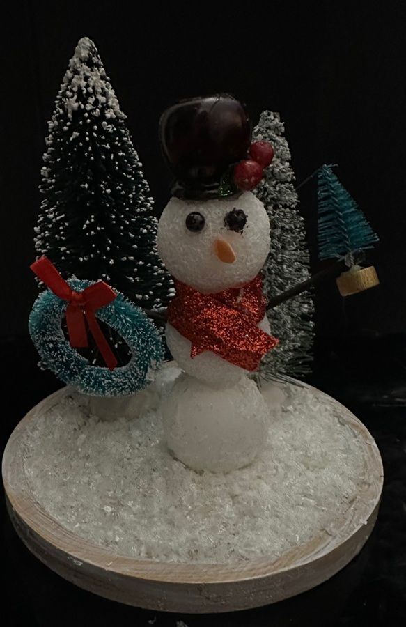 Snowman figurine with black hat, red scarf, and wreath, standing on a white base with miniature trees