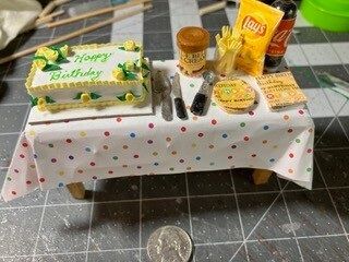 Mini birthday cake with candles, snacks, a drink, and small accessories on a polka-dot tablecloth.