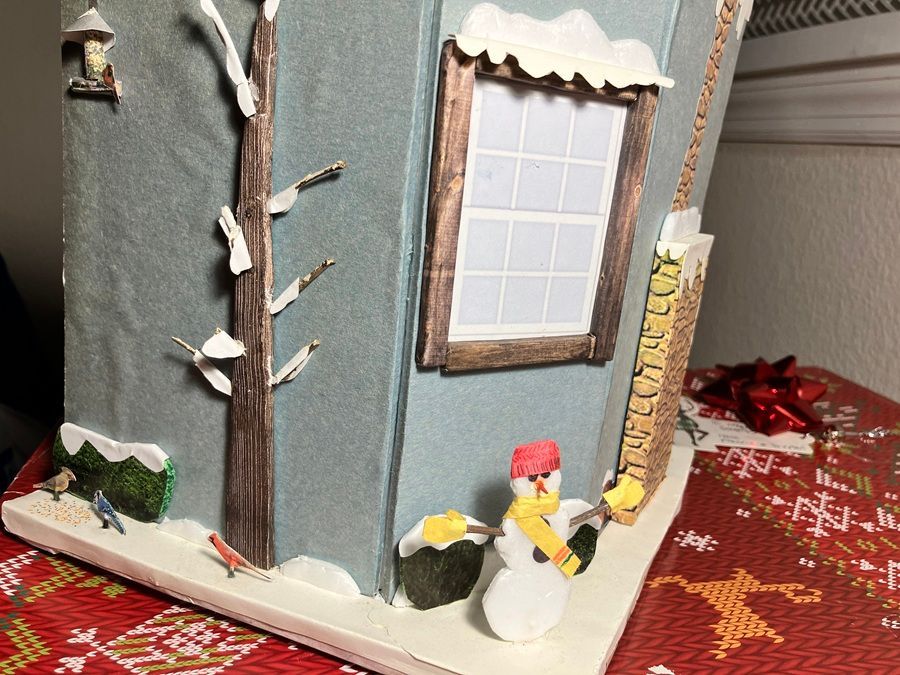 Miniature winter house display with snow, a window, a small snowman, and red decorations on a patterned surface