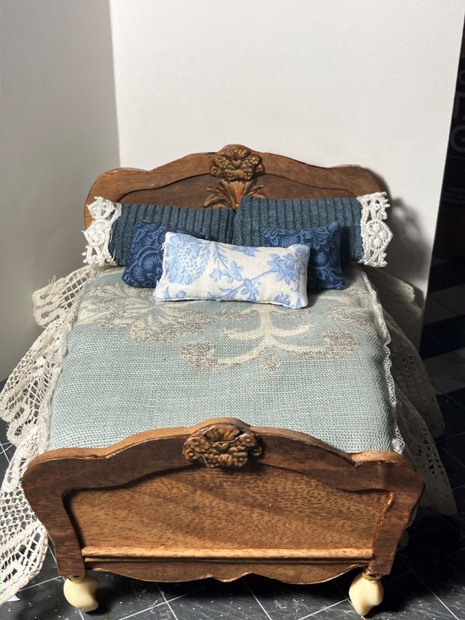Miniature carved wooden bed with blue bedding, lace trim, and pillows against a white wall.