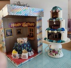Miniature birthday diorama with a "Happy Birthday" booth and a three-tier cake decorated with blue flowers.