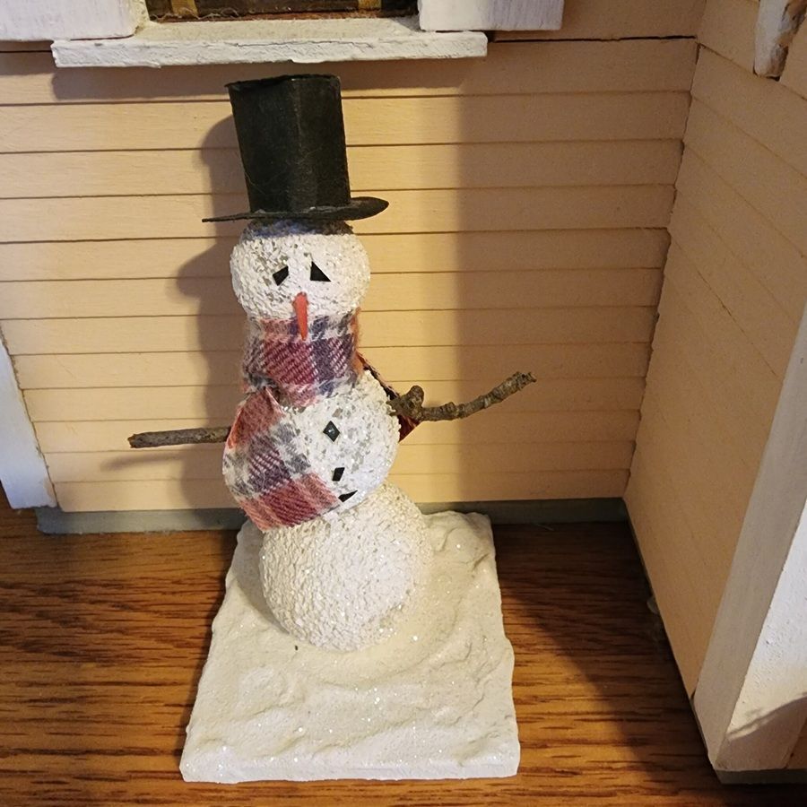Snowman figurine with black top hat and plaid scarf, standing indoors by a wall and doorway.
