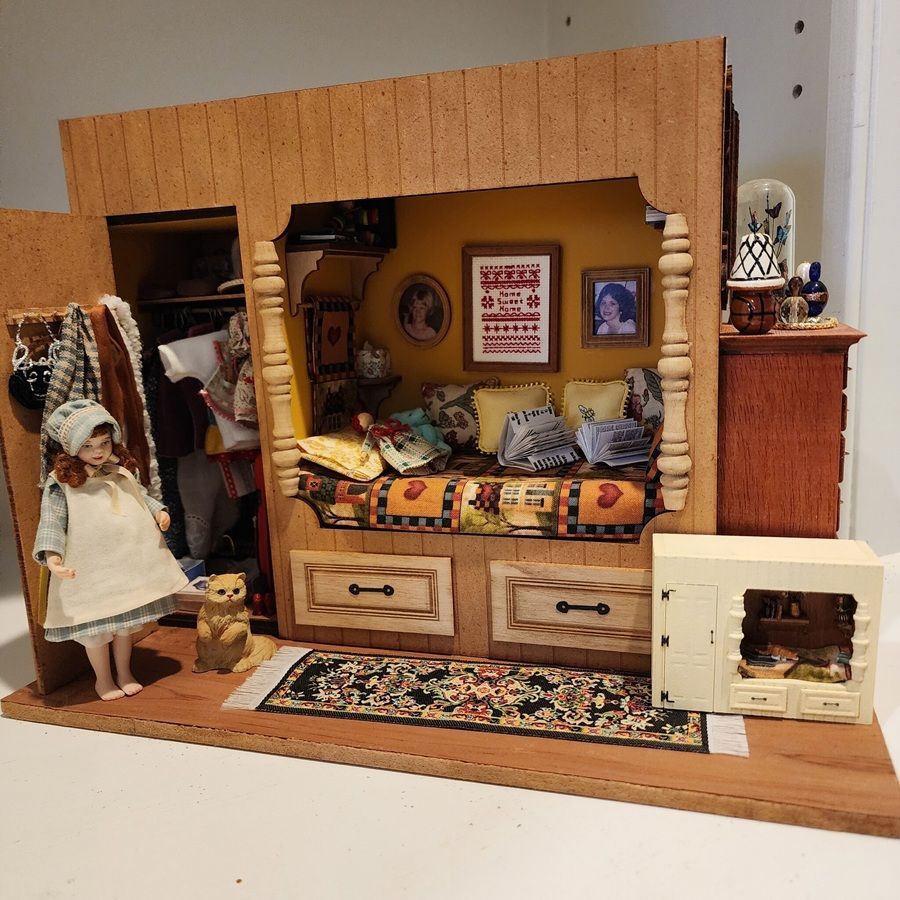 Miniature dollhouse bedroom with bed, chairs, dresser, rug, and a doll in a wooden room display.