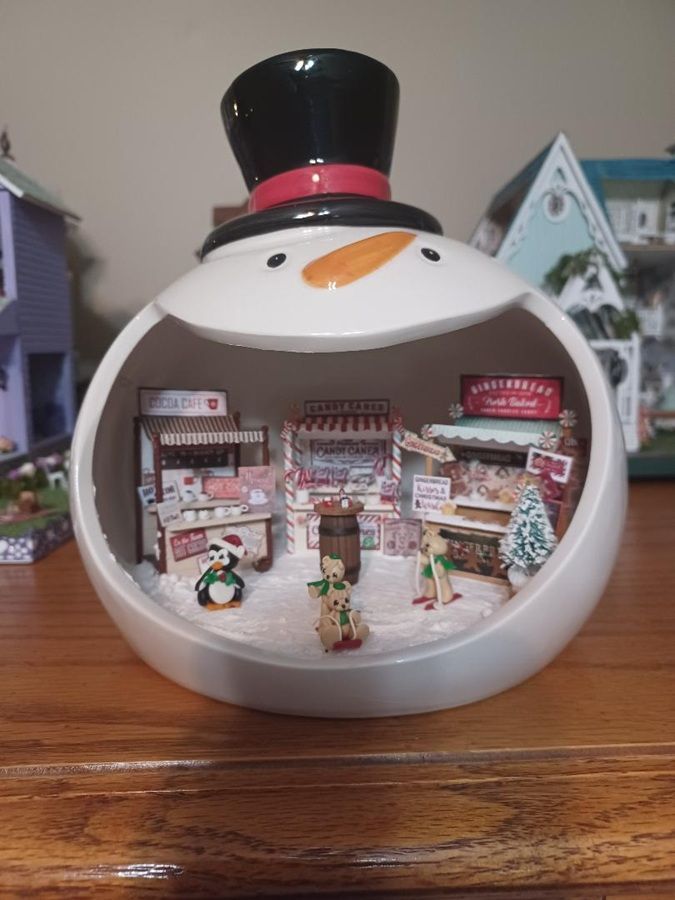 Snowman-shaped display with miniature holiday village scene inside, on a wooden table.