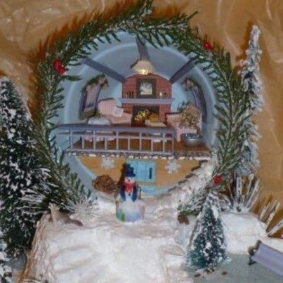 Snowy Christmas village scene with a wreath-framed toy house and tiny pine trees