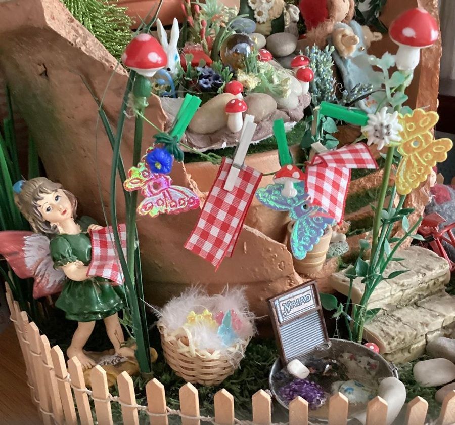 Miniature fairy garden with a doll, striped flags, mushrooms, and flowers behind a small picket fence