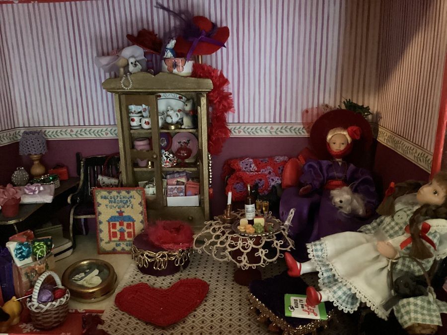 Cluttered doll display with teddy bears, dolls, and miniature furniture in a pink room
