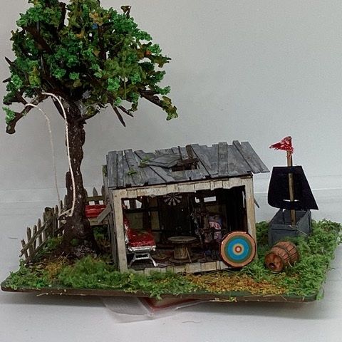 Miniature rustic shed scene with tree, fence, target, barrels, and small black structure on grass.