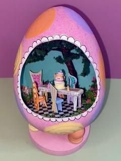 Pink egg-shaped toy open to a miniature outdoor scene with a table, chair, and dessert.