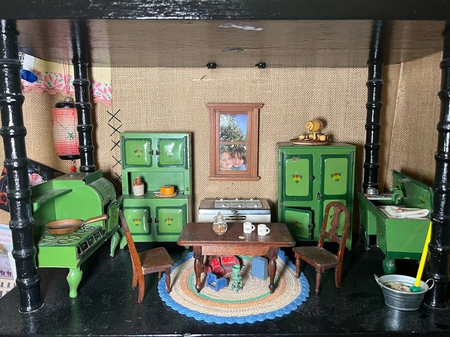 Miniature green dollhouse room with furniture, table, cabinets, stove, and woven rug beneath a shelf structure