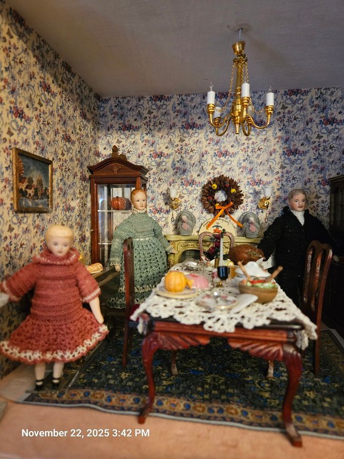 Miniature Victorian-style parlor with dolls around a dining table, floral wallpaper, and a chandelier.