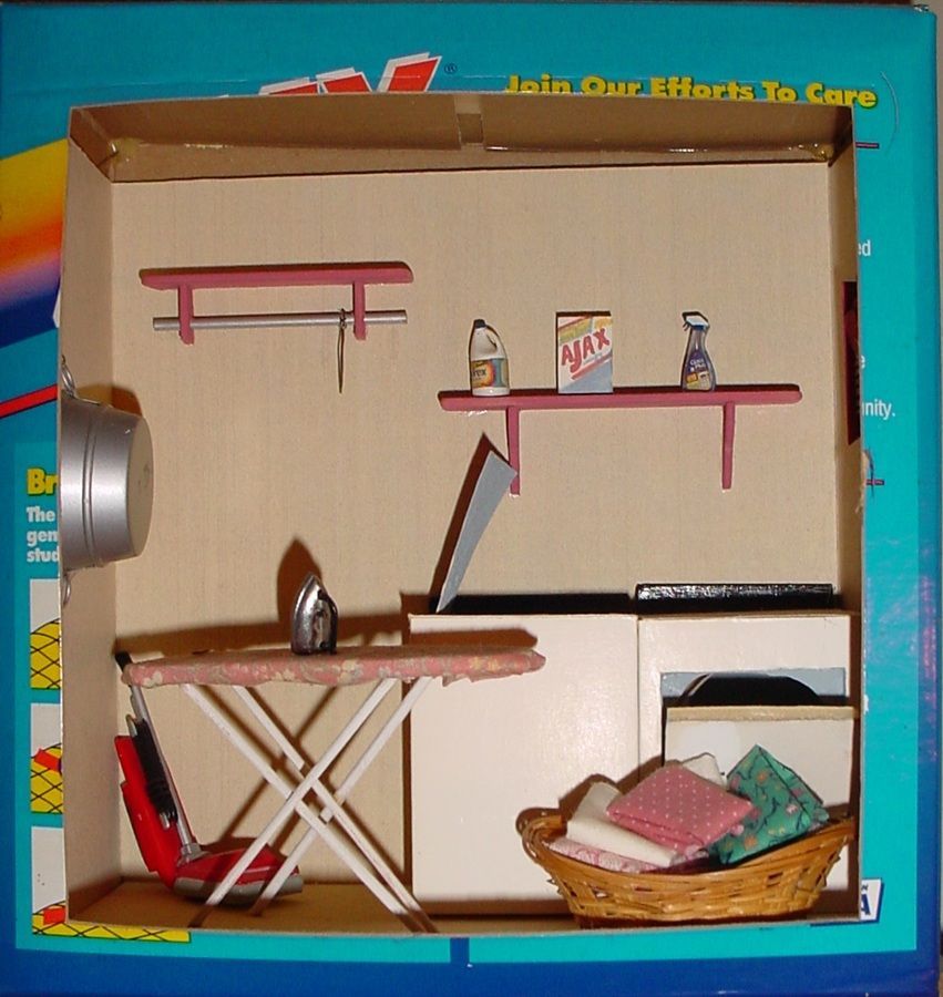 Cardboard dollhouse interior with shelves, tiny table, ironing board, basket, and miniature household items.