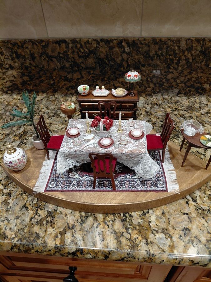 Miniature dining room display with red chairs, patterned rug, and decorative table setting on a granite counter