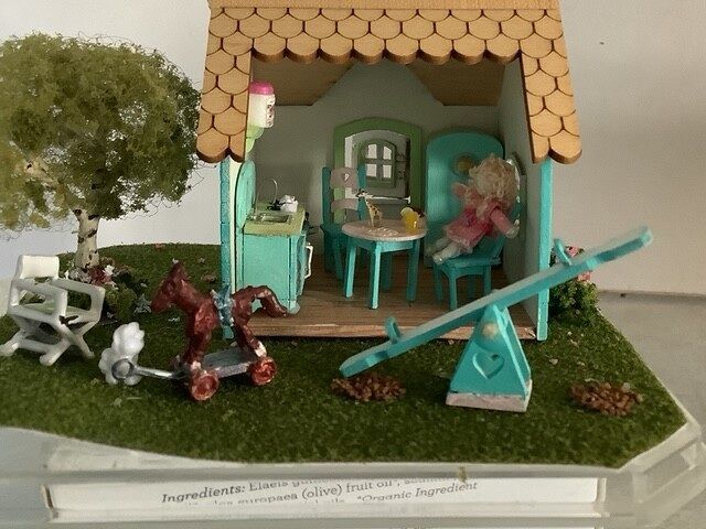Miniature dollhouse scene with green yard, orange roof, table, pink chair, and a seesaw in front