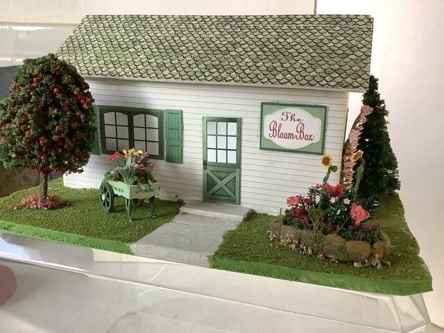 Miniature white house with green roof, flowers, and a sign reading “The Flower House”