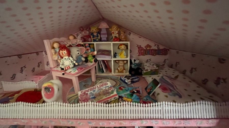Pink dollhouse attic room filled with toys, miniature shelves, and colorful dolls on a sloped ceiling