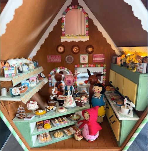 Miniature bakery display in a dollhouse, with pastries, shelves, and two dolls inside a cozy shop setting