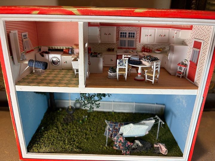Dollhouse interior with kitchen, laundry room, and small bedroom scene with a bed and greenery.