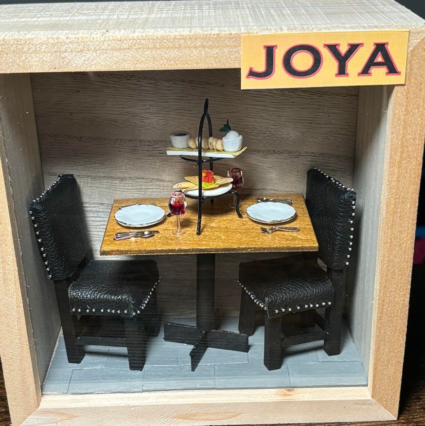 Miniature restaurant booth with two black chairs, a wooden table, and a JOYA sign above.