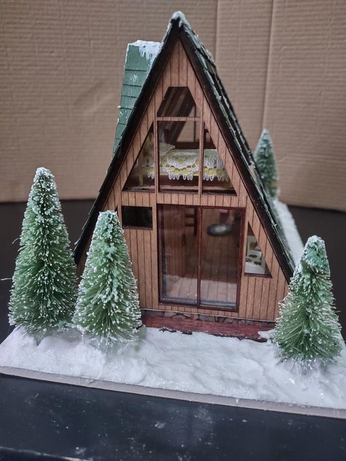 Miniature A-frame cabin with frosted evergreen trees on a snowy base and black roof trim
