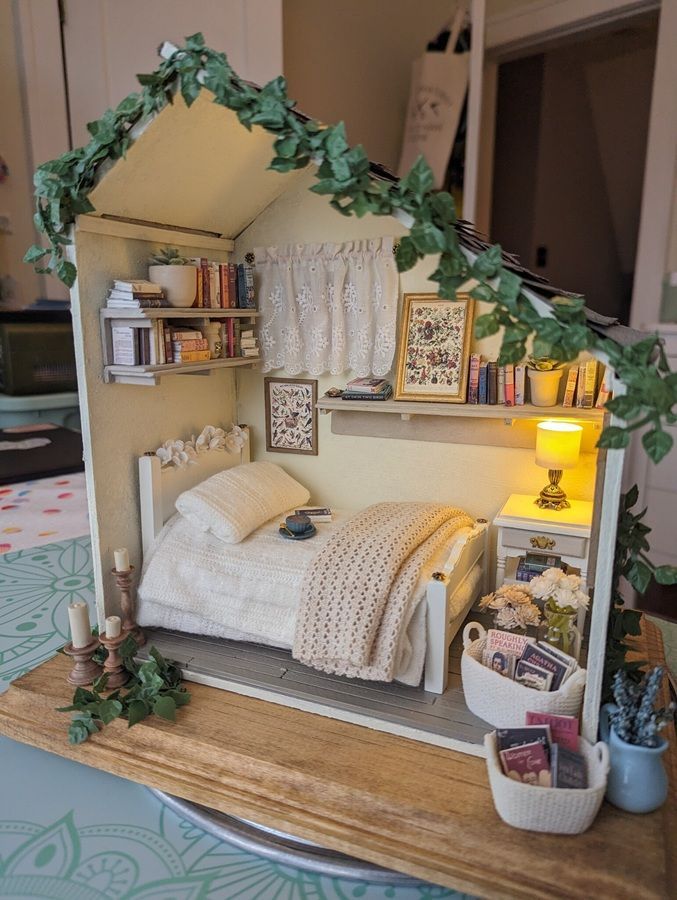 Miniature bedroom diorama with white bed, wooden floor, leafy arch, shelves, and warm bedside lamp