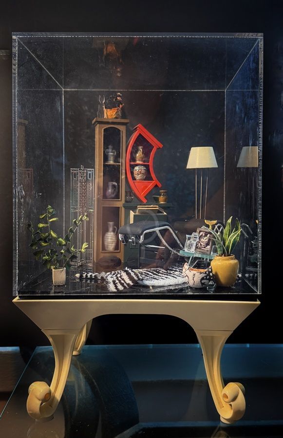Glass display case with a small room vignette, gold stand, plants, chair, and lamp inside