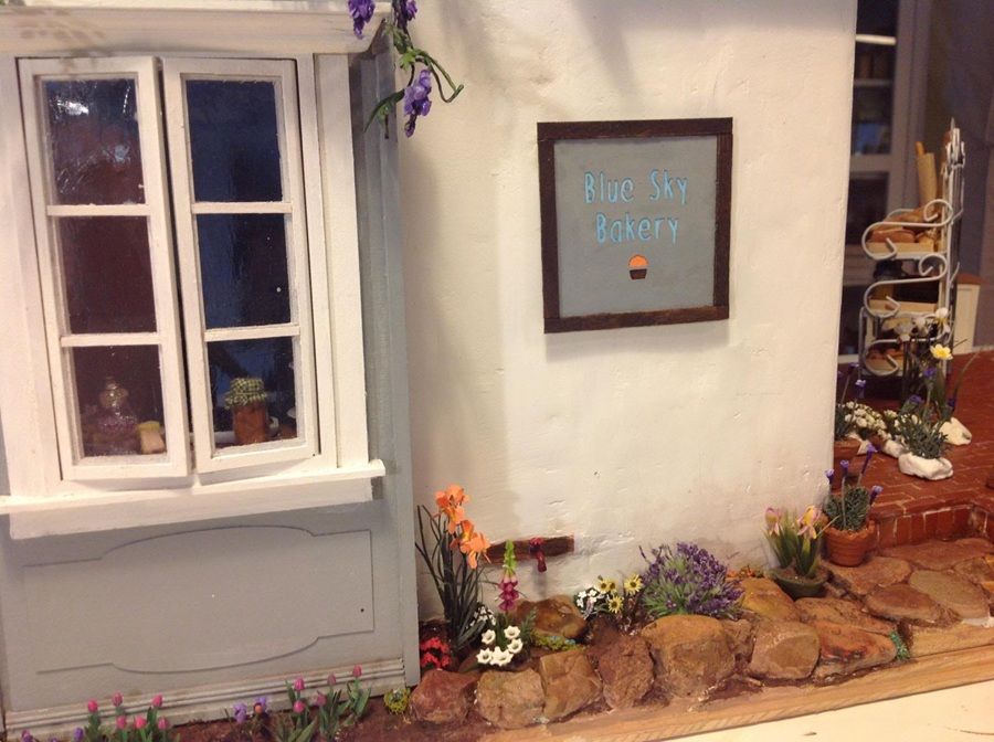 White cottage wall with window and framed sign, flower pots, and a small stone-bordered garden path.
