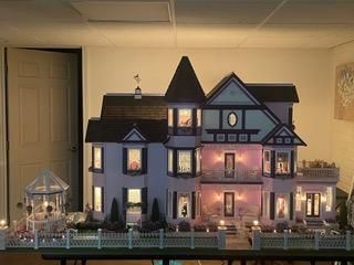Miniature Victorian-style house model with lit windows, a gazebo, and a front fence on a shelf indoors