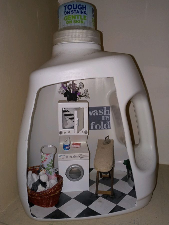 Laundry room diorama inside a detergent bottle, with a mini washer, basket, and chair on a tiled floor.