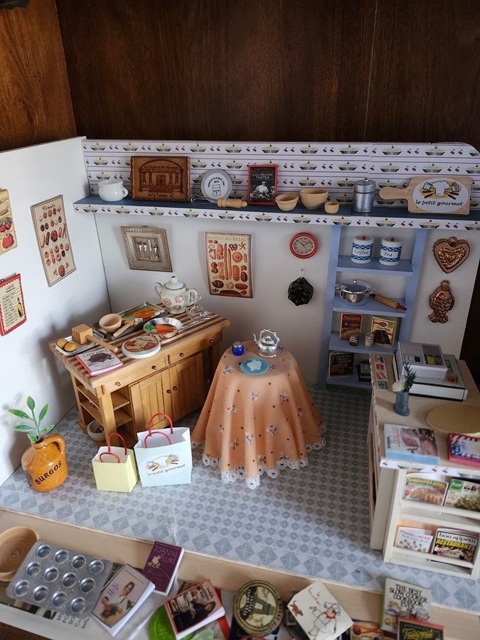 Miniature dollhouse room with tiled floor, wooden cabinets, round table, and shelves filled with tiny household items.