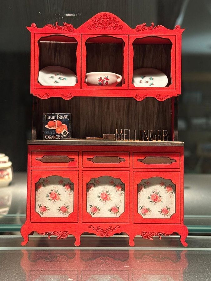 Red wooden doll cabinet with floral panels and shelves holding white dishes, centered against a dark background