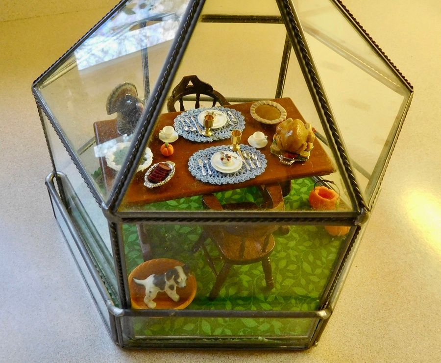 Mini glass terrarium with a small dining room scene, table set with plates and pastries on green flooring.