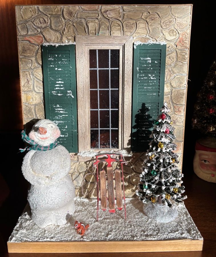 Snowman and Christmas tree in front of a house façade with green shutters and a central door.