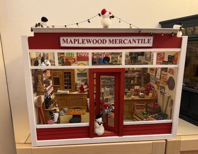 Miniature storefront labeled “Maplewood Mercantile” with red trim and holiday decorations inside a display case
