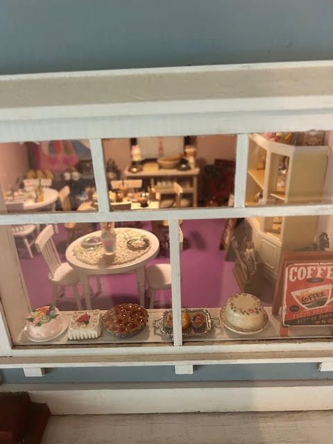 Miniature café display seen through a window, with pastries, a round table, and a red coffee sign.