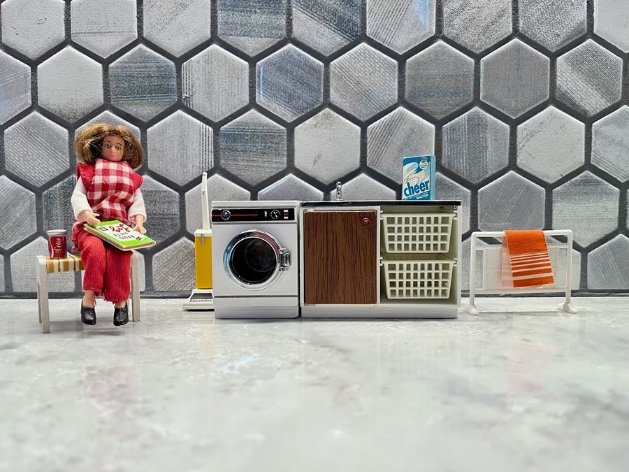 Miniature living room scene with a doll, washer, cabinet, and sofa against a hexagon-pattern wall