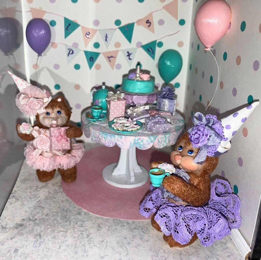 Two teddy bears in purple and pink dresses at a pastel tea party with balloons and a cake table