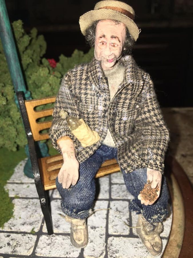 Small figurine of a man sitting on a bench, wearing a straw hat, plaid coat, jeans, and work boots.