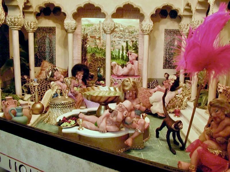 Miniature ornate palace scene with doll-like figures, gold decor, and bright pink feathers