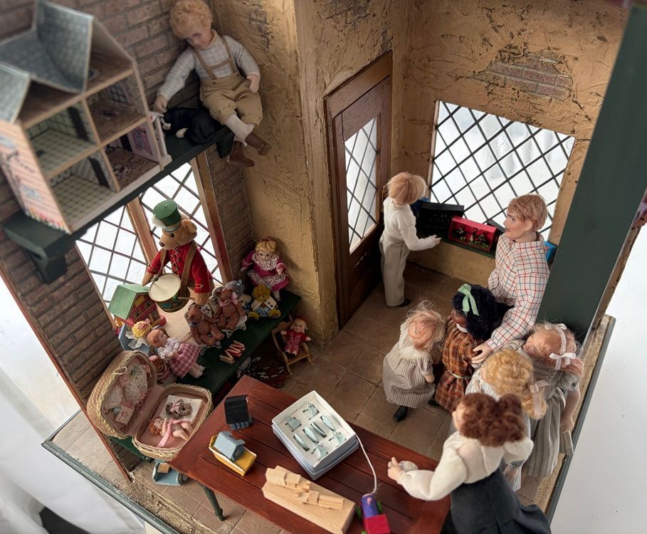 Miniature room scene with several figures gathered around a desk, shelves, windows, and plants.