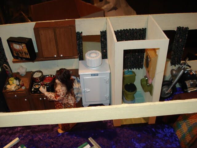 Miniature dollhouse kitchen with cabinets, fridge, sink, and a seated doll on the left.