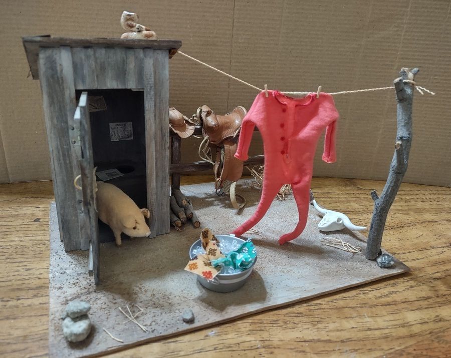Small diorama with a red hanging suit, toy animals, and a tiny figure around a rustic shed.