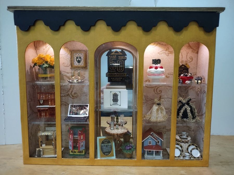 Miniature display cabinet with arched shelves holding tiny decor, cakes, flowers, books, and a dollhouse