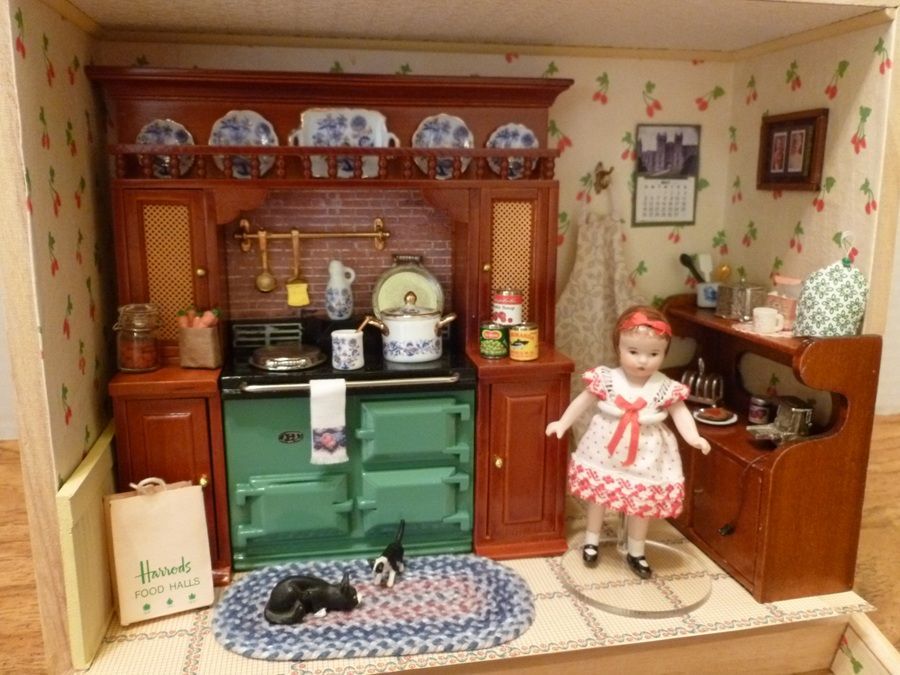 Miniature vintage kitchen scene with green stove, wooden cabinets, doll on the right, and a black cat on the floor