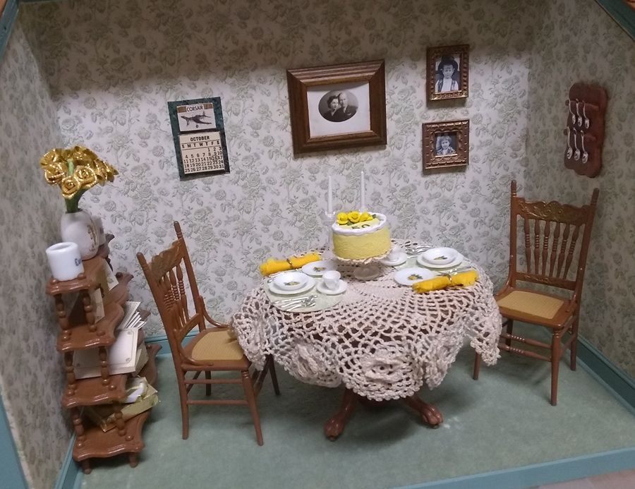 Miniature dollhouse dining room with lace-covered table, tea set, chairs, and framed wall art