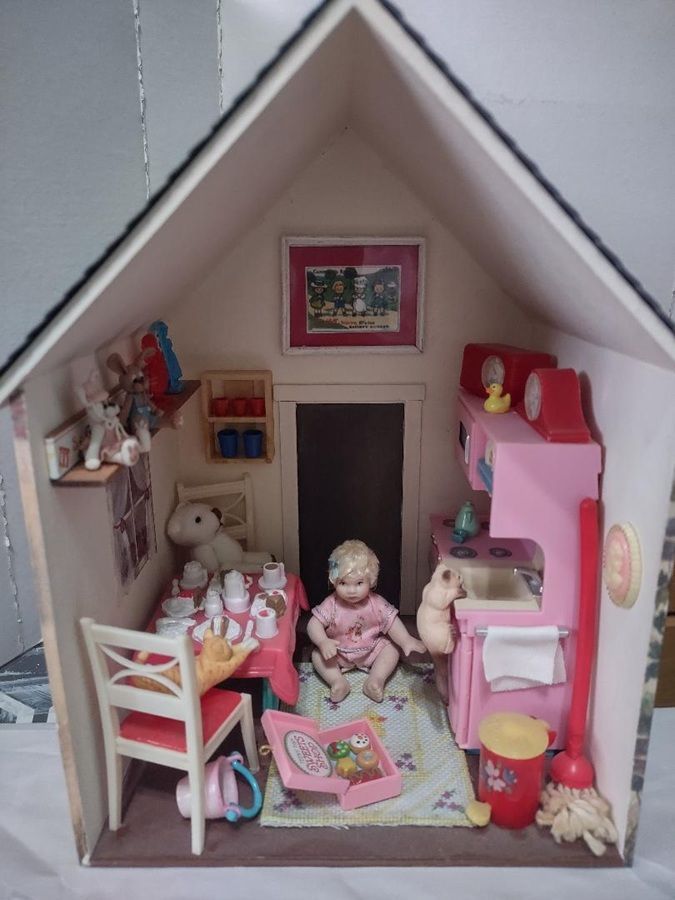 Miniature dollhouse room with pink furniture, dolls, and a central doorway under a peaked roof.