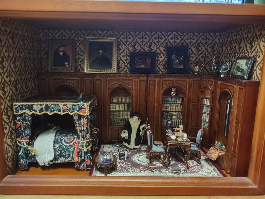 Miniature room display with ornate wood walls, a canopy bed, seated figures, and framed portraits above.