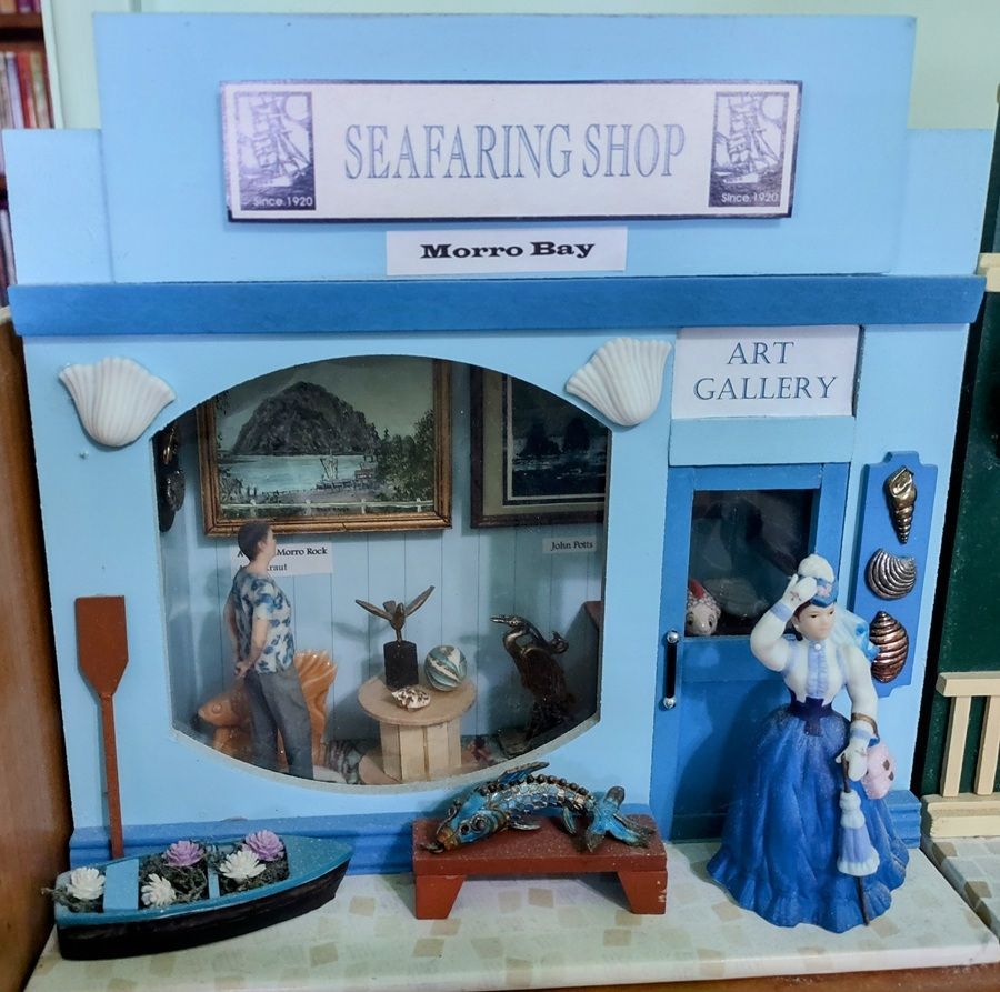 Blue seafaring shop miniature with art gallery, boat display, and sailor figurine in front of a storefront