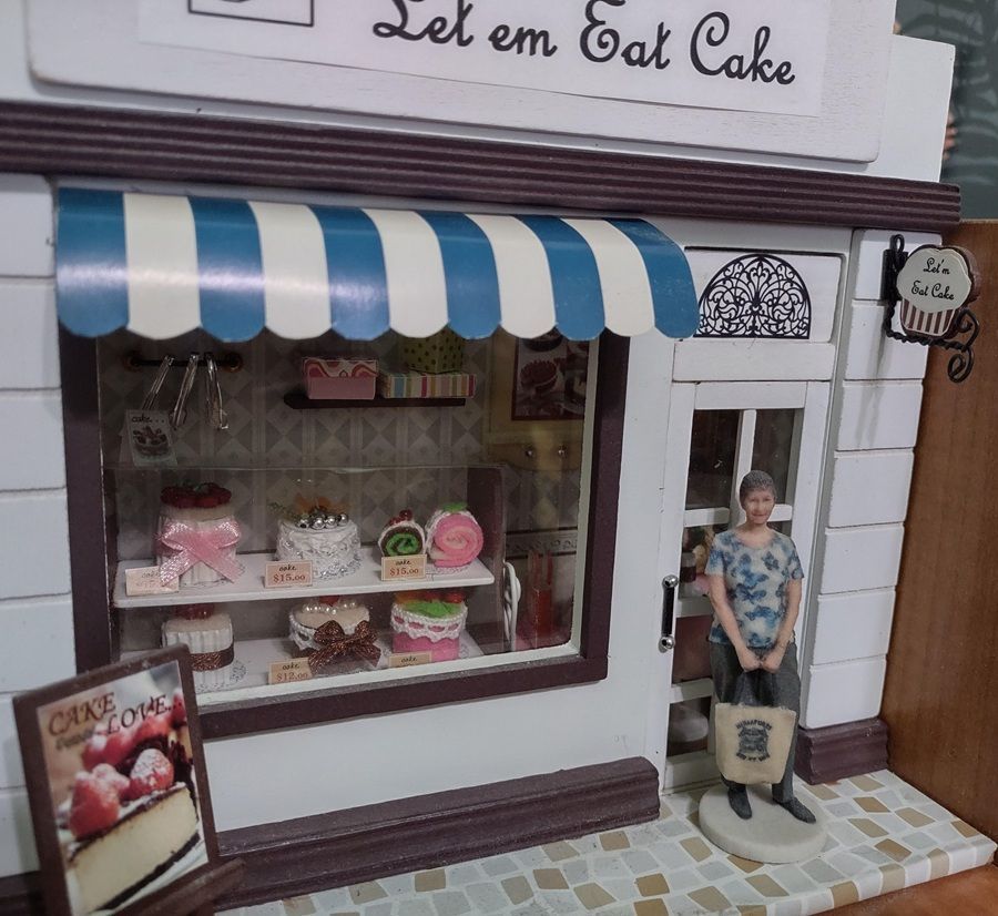Miniature cake shop façade with blue-striped awning, pink cakes in the window, and a small figure standing outside.