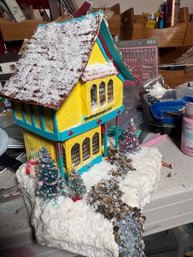 Colorful miniature house with snow-covered roof on a craft table, surrounded by winter decorations